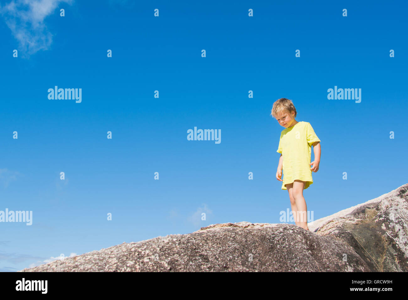 A Blond Boy In A Yellow T-Shirt Is Standing On A Rock In Front Of Blue Sky Stock Photo