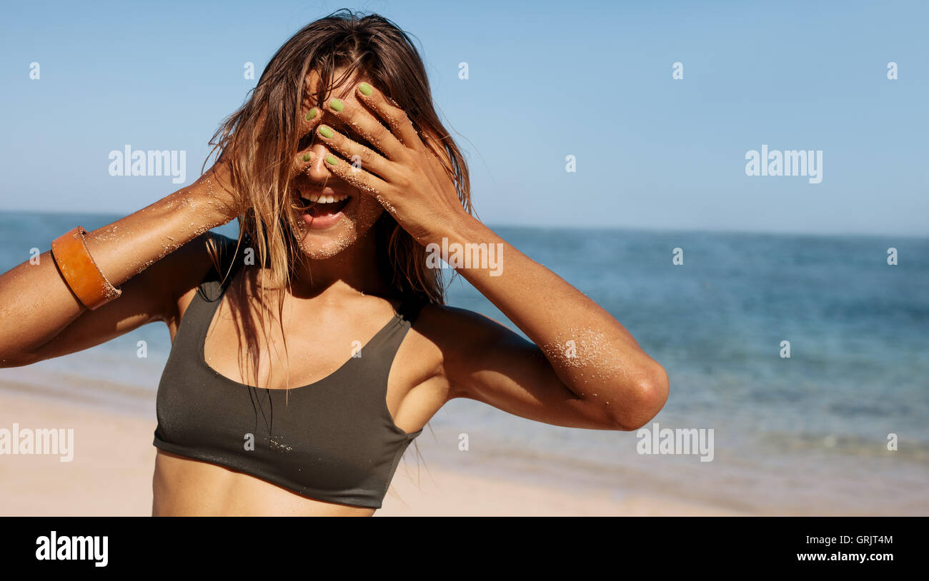 Portrait of happy young woman standing outdoors on the beach and cover her eyes with hands. Female bikini model on sea shore hav Stock Photo