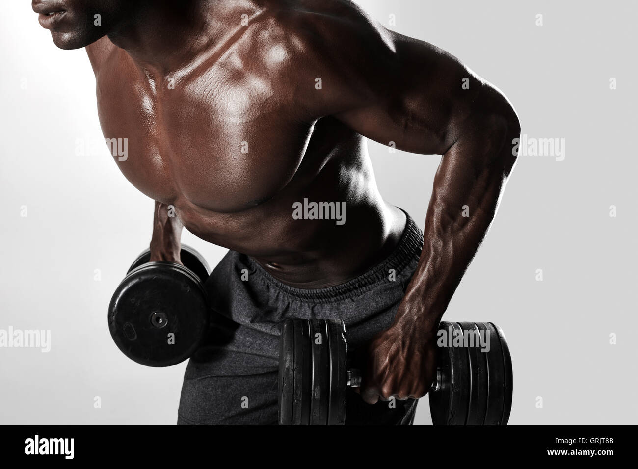 Close up shot of african bodybuilder exercising with dumbbells. Young man doing exercise with heavy dumbbells over grey backgrou Stock Photo