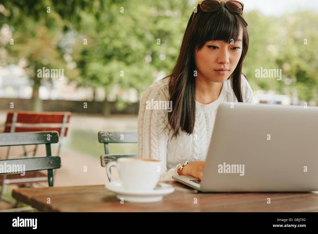 Shot of young woman sitting at cafe table outdoors and working on laptop computer. Asian female surfing internet on laptop. Stock Photo