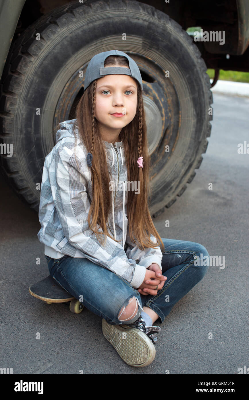 girl sit on skateboard outdoor near huge rusty truck wheel Stock Photo