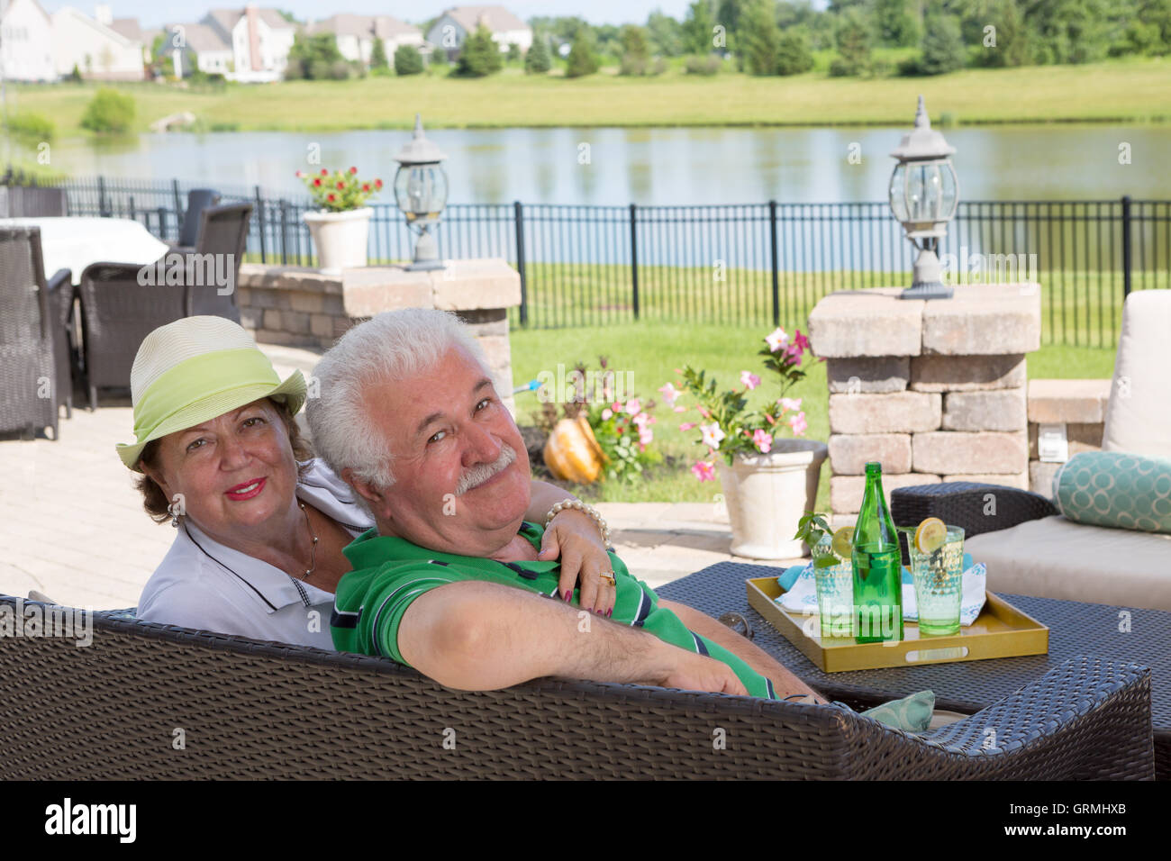 Affectionate elderly couple enjoy a relaxing day on the patio sitting close together on a wicker settee overlooking a scenic pon Stock Photo