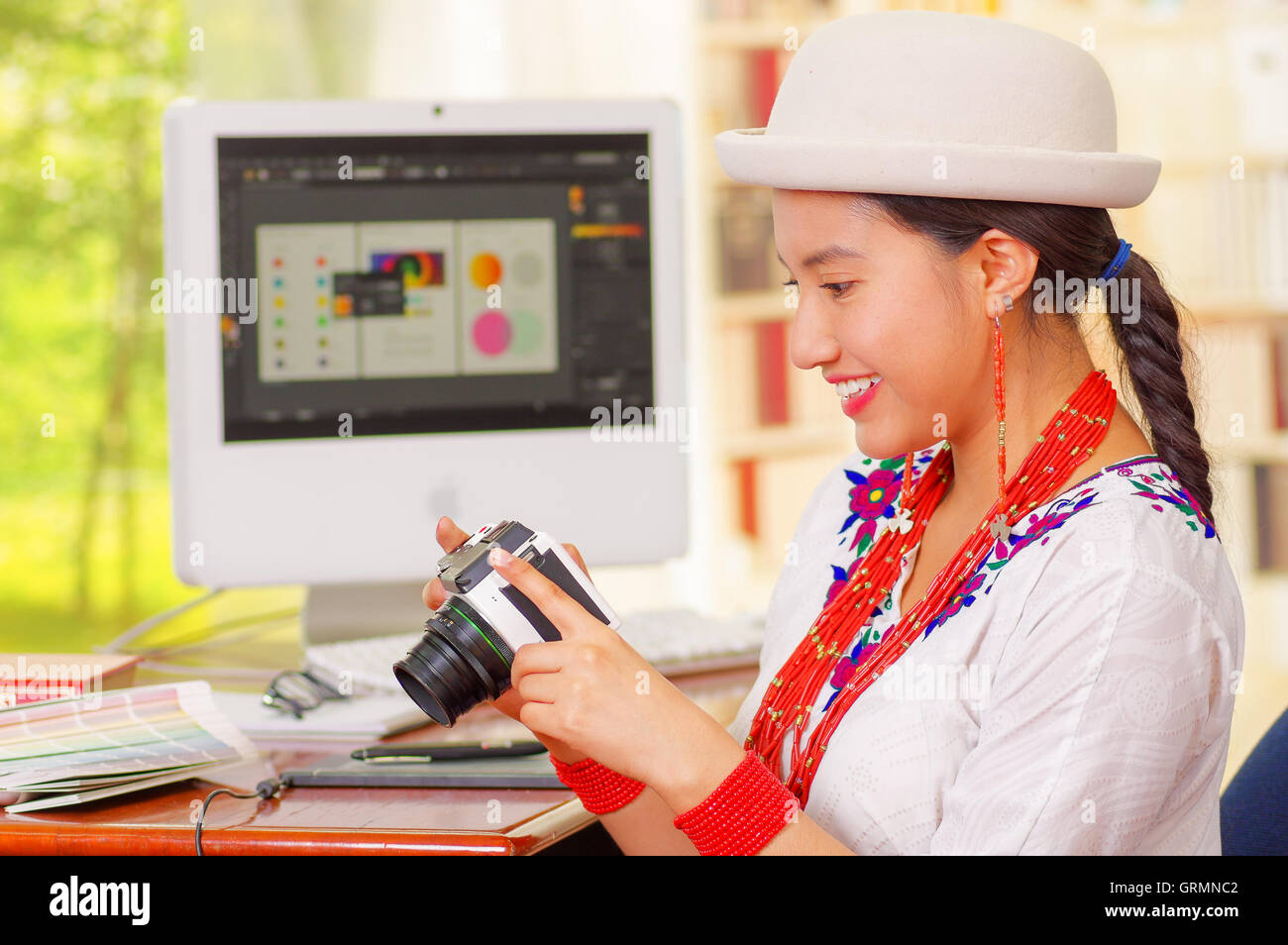 Young pretty girl wearing white shirt and fashionable hat, sitting by computer desk holding a camera, seen from profile angle Stock Photo