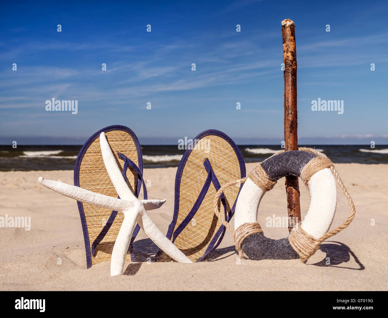 Life buoy, starfish and pair of straw flip-flops stuck into the sand against the sea and sky Stock Photo