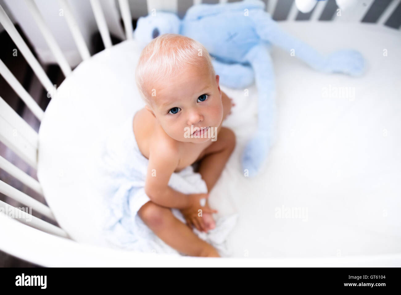 Cute baby sitting in a white round bed. White nursery for young children. Little boy crying in his crib. Toys for infant cot. Stock Photo