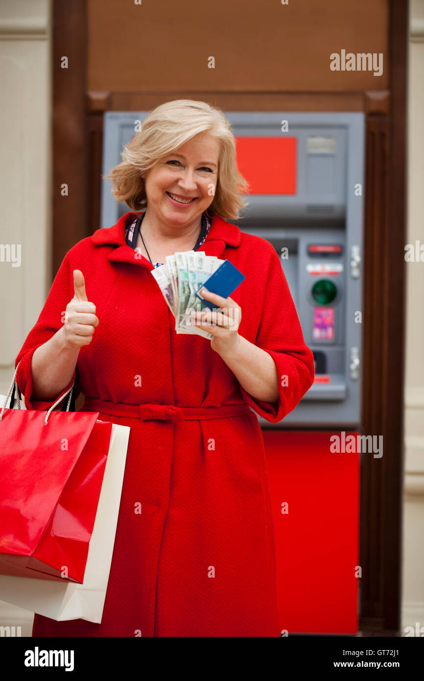 Mature blonde woman with shopping bags on the background of the ATM Stock Photo