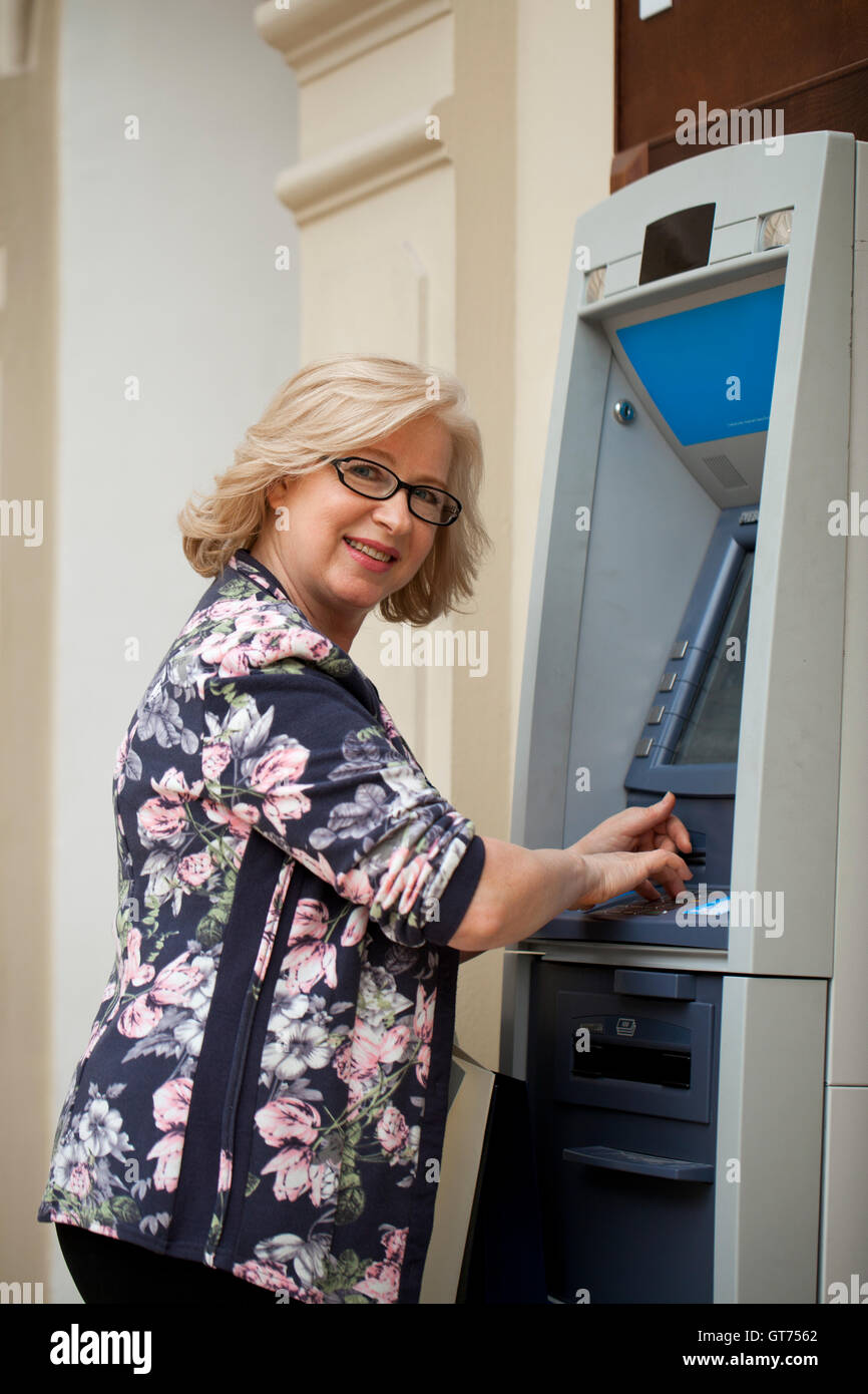 Mature blonde woman counting money near automated teller machine in shop Stock Photo