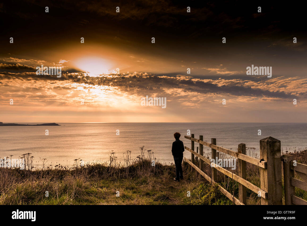 A woman stands on cliffs looking at a stunning sunset breaking over Newquay Bay in Cornwall. Stock Photo
