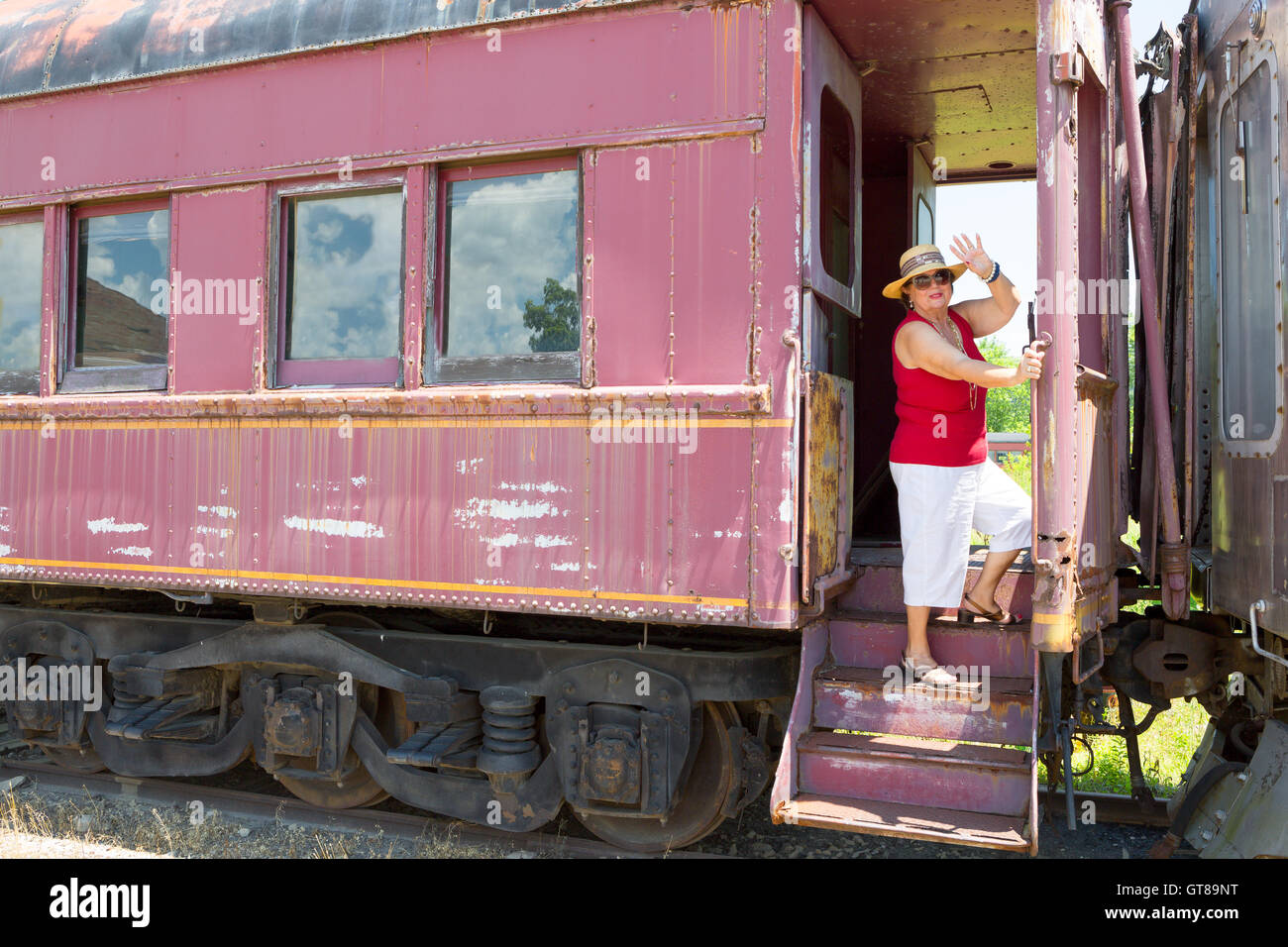 Elderly woman in trendy summer clothing standing waving at the camera from the steps of a vintage train carriage in a conceptual Stock Photo