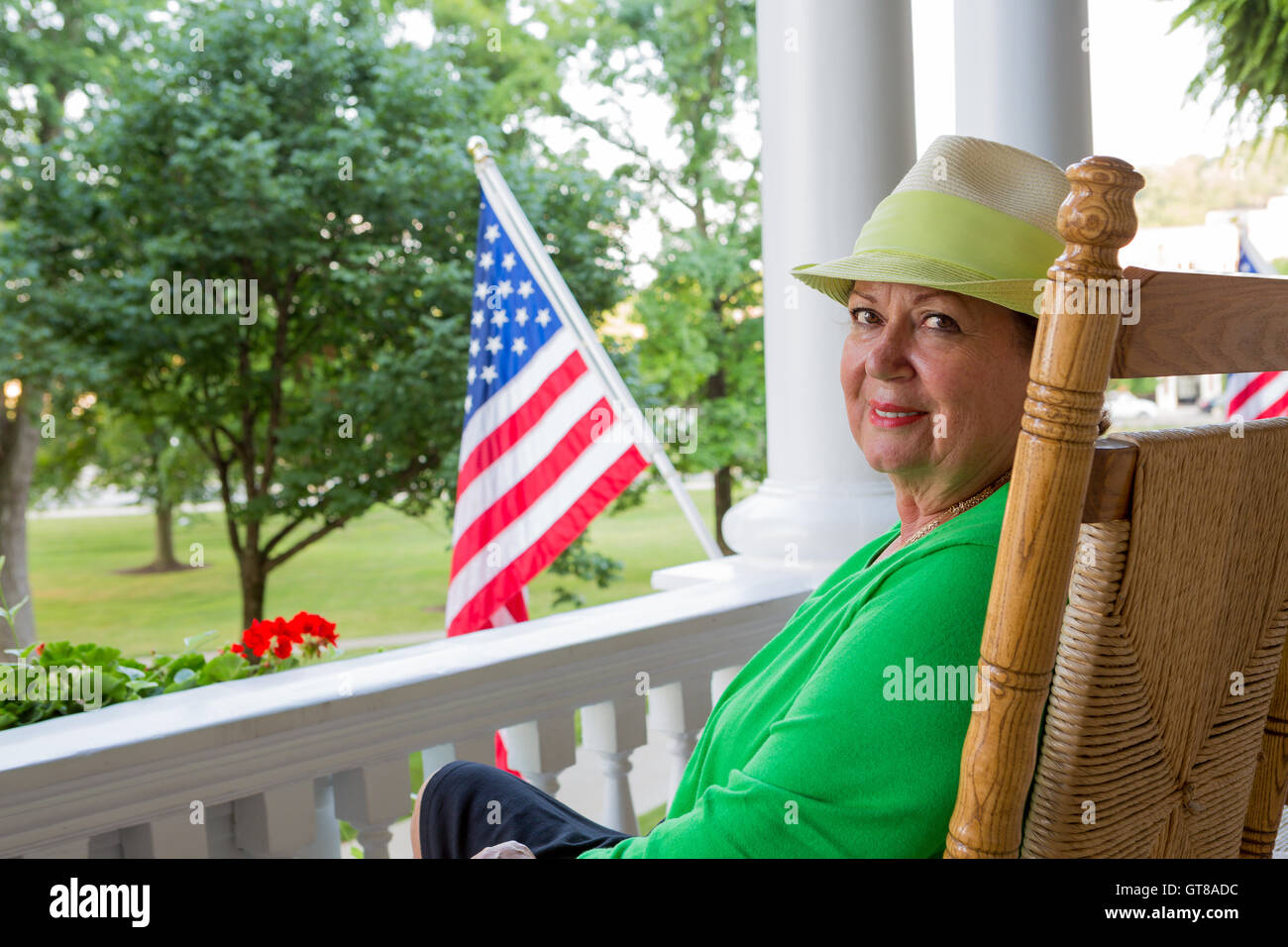 Attractive friendly trendy elderly lady in a green top and hat sitting on her patio with the American national flag fying alongs Stock Photo