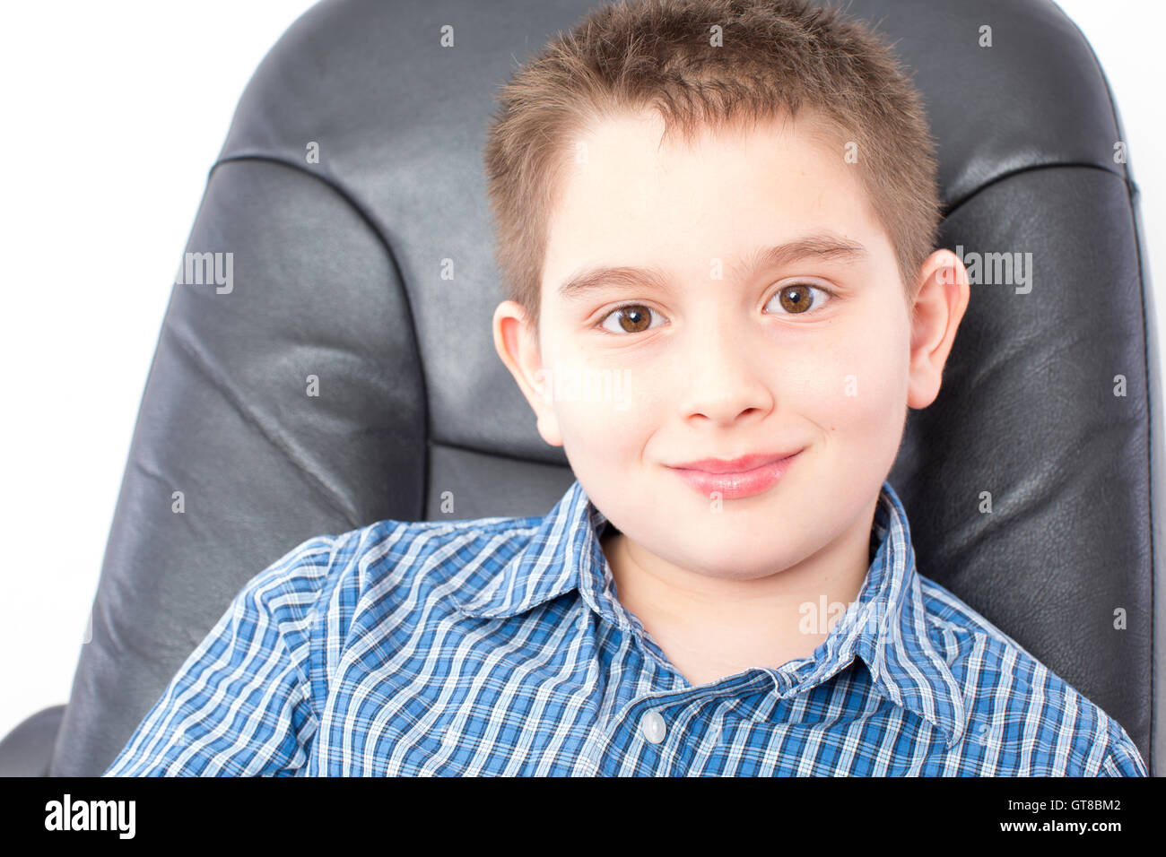 Close up Smiling Cute American Boy Sitting on a Black Office Chair, Looking at the Camera, on a White Background. Stock Photo