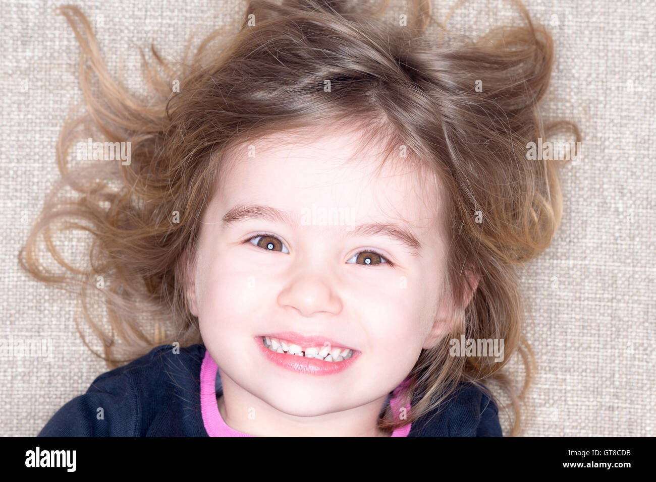 Headshot of young three year old girl lying on a carpet with tousled hair grinning up at the camera with a happy expression Stock Photo