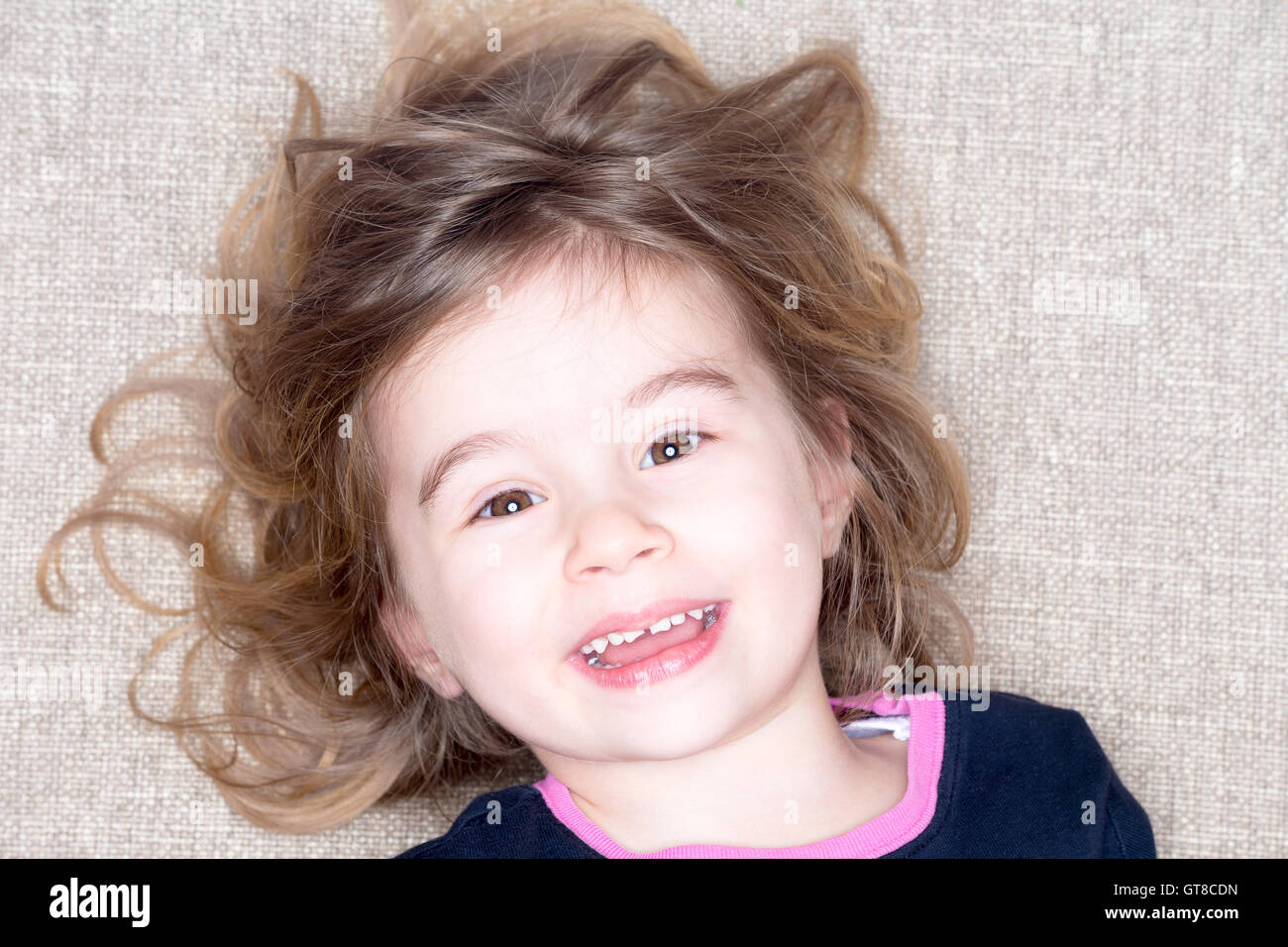 Pretty vivacious three year old little girl lying on a carpet with her hair flying around her face smiling happily up at the cam Stock Photo