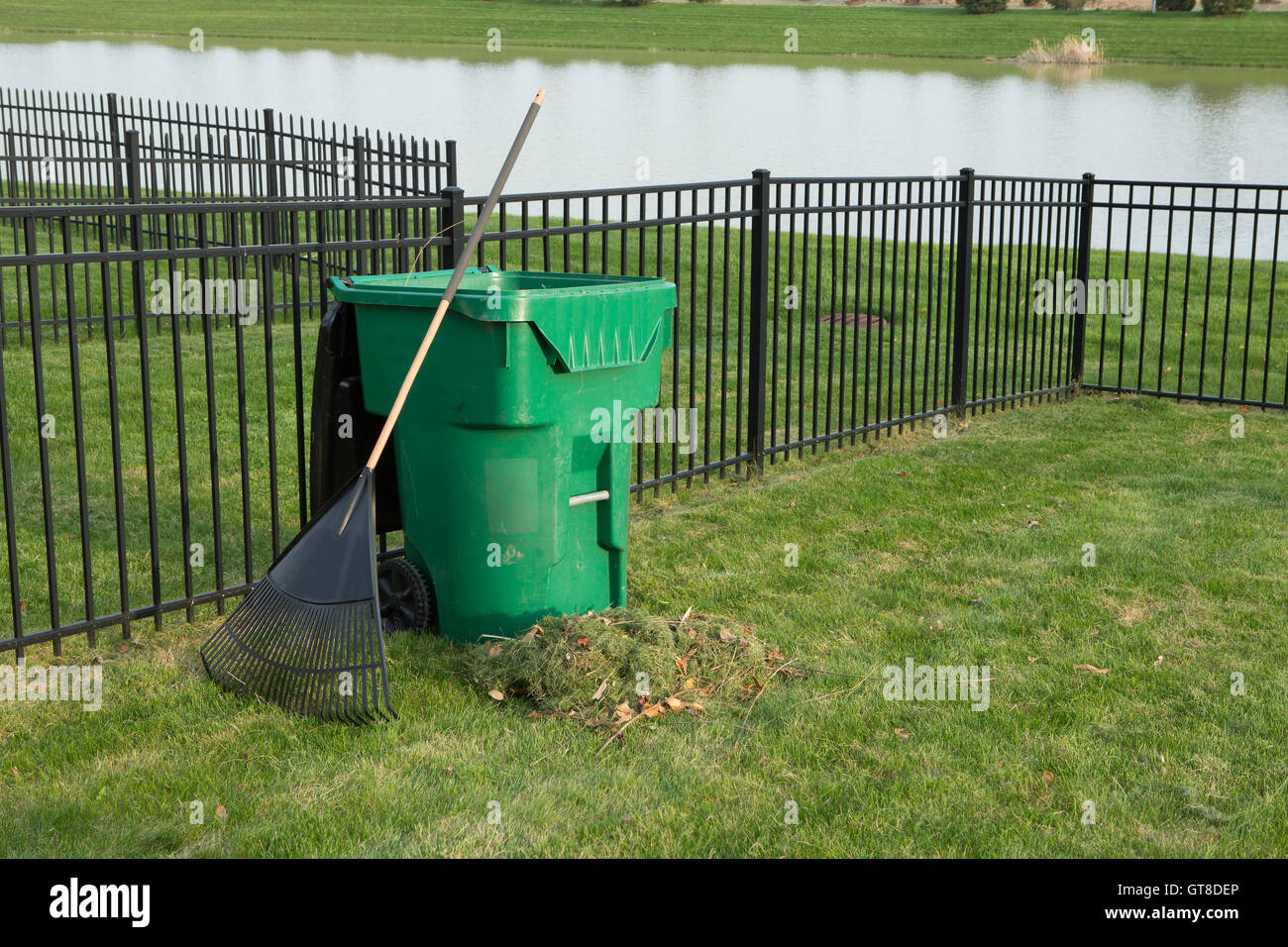 Yard maintenance in spring with a fresh heap of grass clippings and a rake leaning on a green plastic bin for composting organic Stock Photo