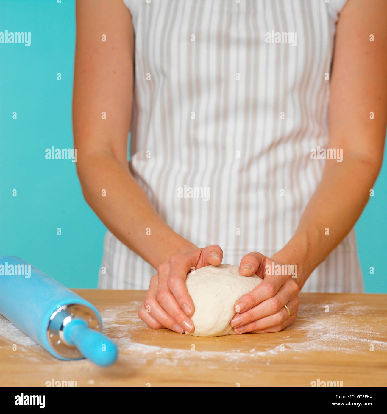 Woman in Striped Apron Kneading Dough on Wooden Surface with Blue Rolling Pin Stock Photo