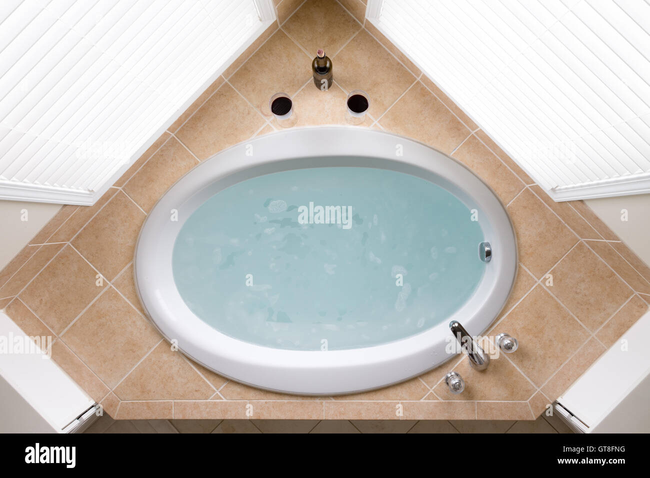 Stylish corner oval bathtub filled with fresh clean water in a brown tile surround flanked by two windows, overhead view Stock Photo