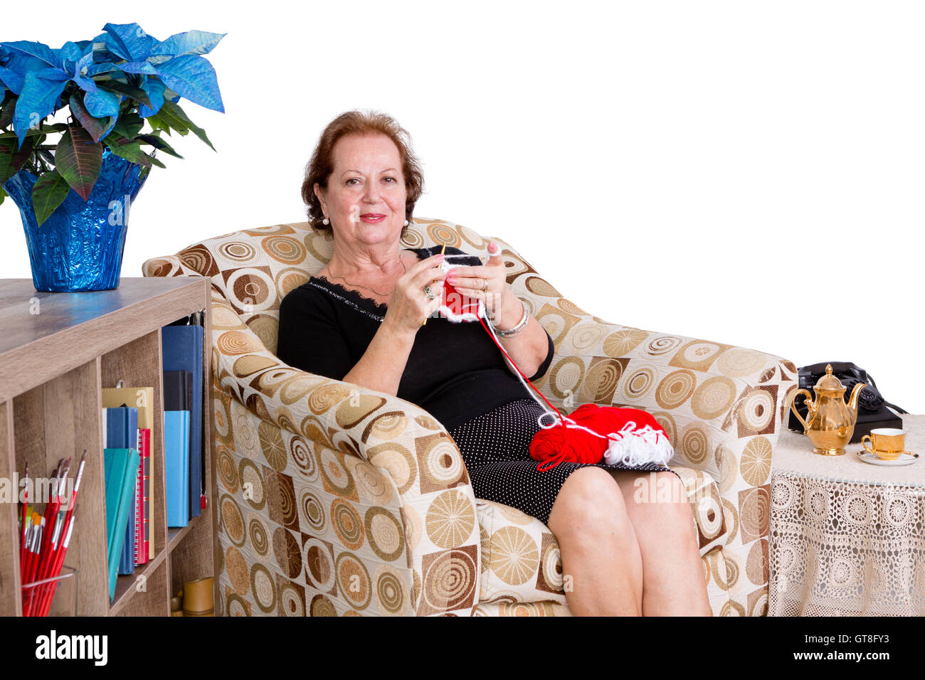 Elegant elderly lady sitting knitting at home in a comfortable armchair looking up to give the camera a friendly smile against a Stock Photo