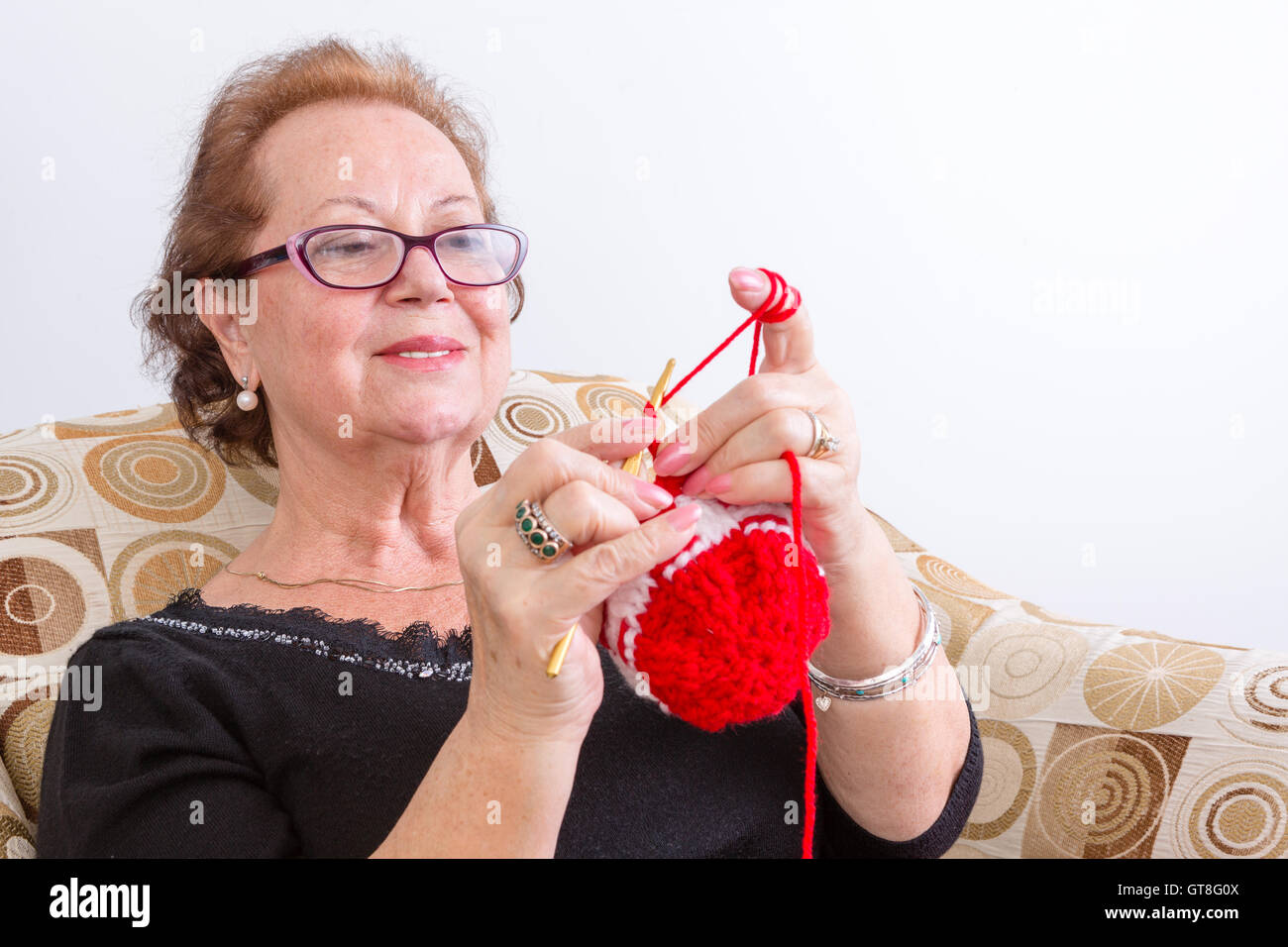 Senior lady sitting knitting at home holding up her colorful red knitting in front of her with a pleased look of concentration, Stock Photo
