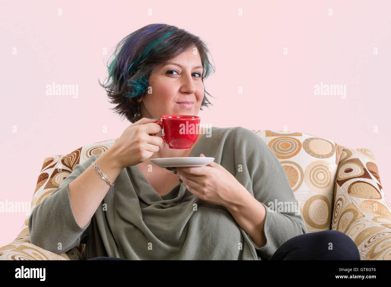 Single intelligent grinning adult female holding red coffee mug while sitting in a comfortable sofa chair Stock Photo