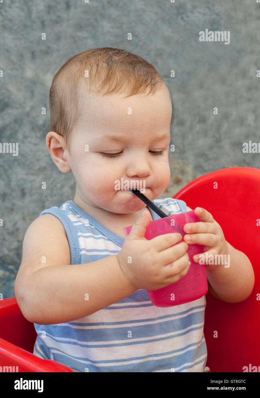Baby boy drinking by himself with straw. He is seated in high chair at terrace restaurant Stock