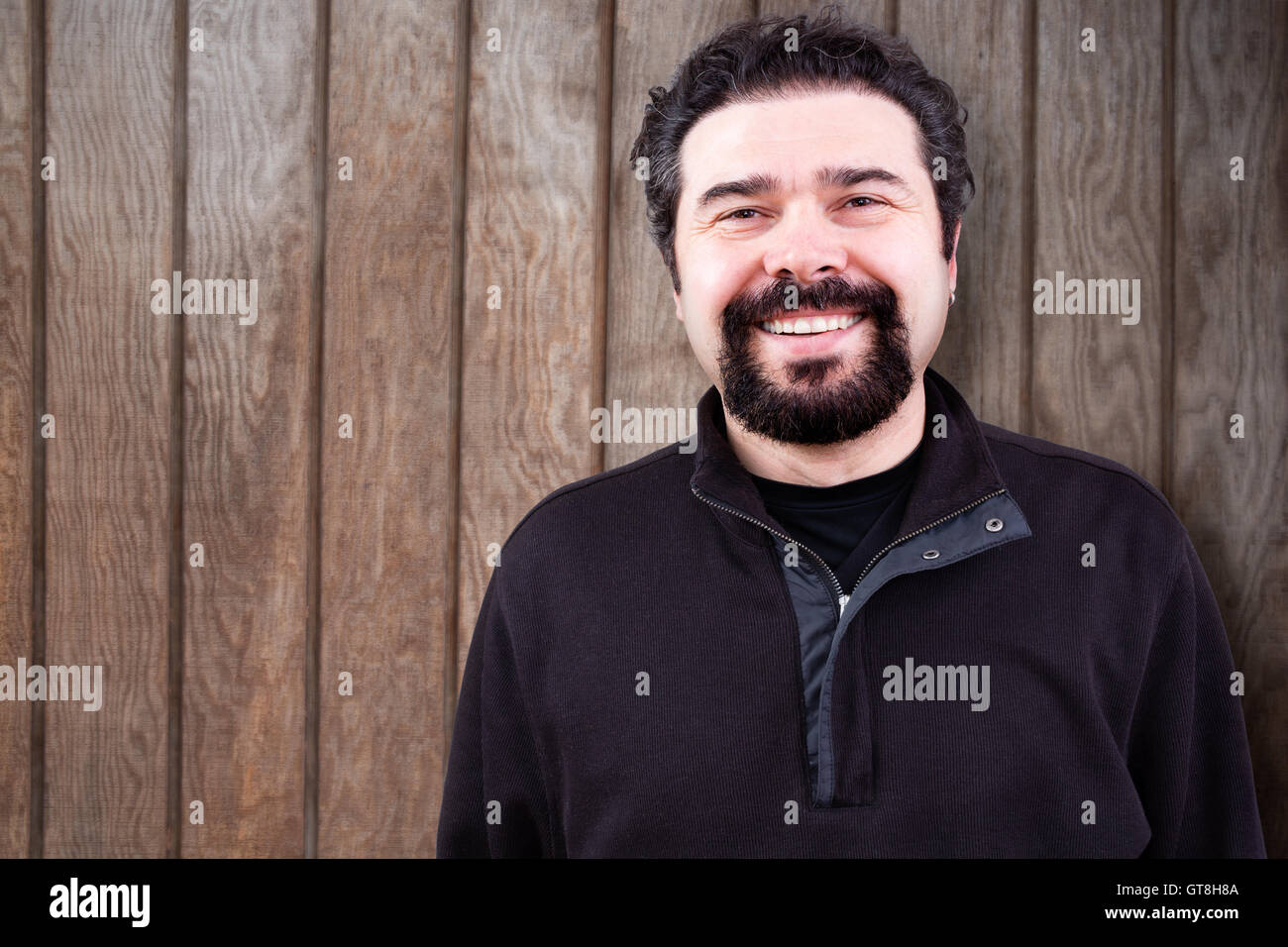 Half Body Shot of a Middle Aged Man with Goatee Beard, Sincerely Smiling at the Camera, Against Wooden Wall with Copy Space. Stock Photo