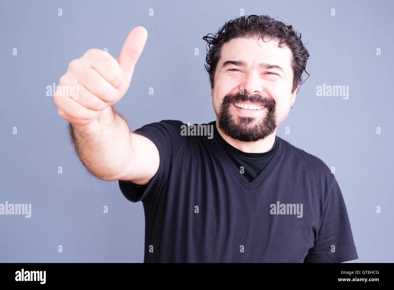 Middle aged bearded single man in black shirt sticking his hand outward with thumb up and big happy smile over gray background Stock Photo