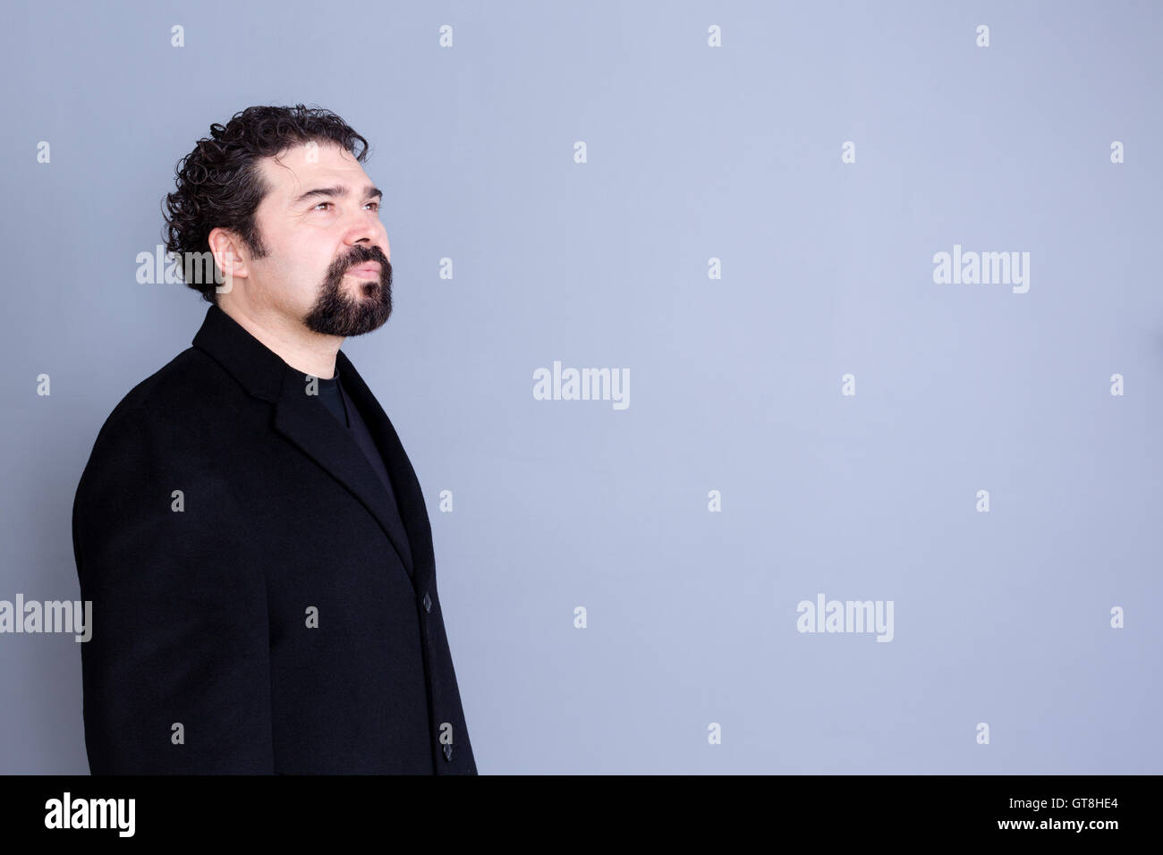 Three quarter view of hopeful attractive dark haired and bearded middle aged male in black shirt and blazer looking away over gr Stock Photo