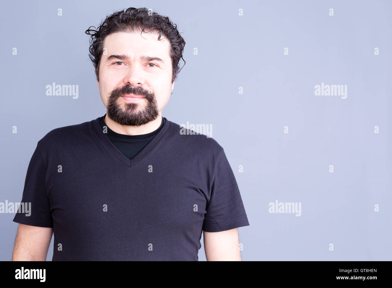 Waist Up Portrait of Mature Man with Beard and Dark Curly Hair Wearing Black T-Shirt and Staring at Camera with Blank Contented Stock Photo