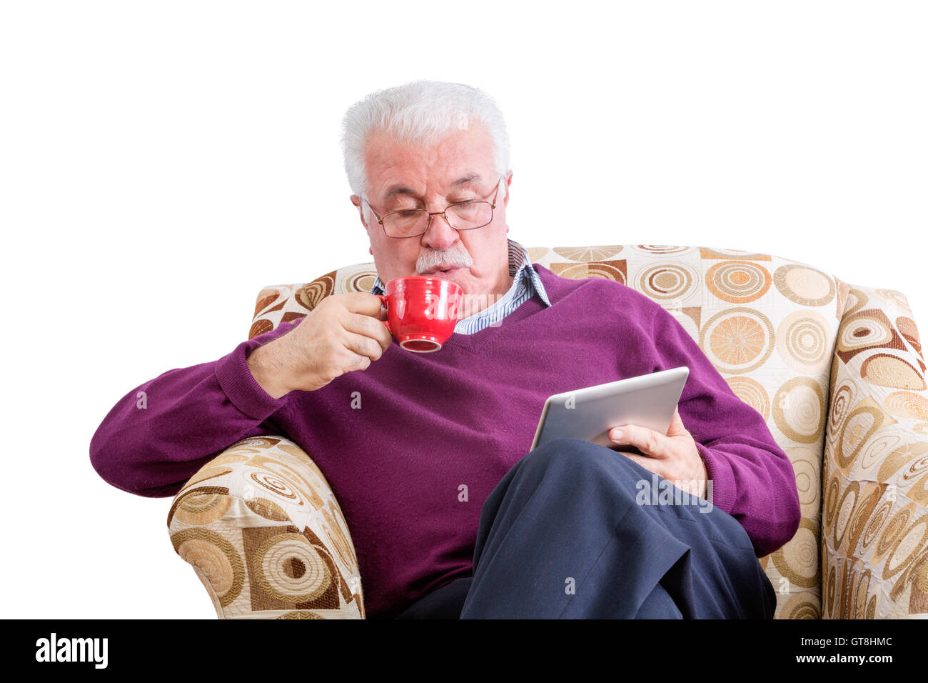 Senior male in purple sweater with mustache looking down at tablet computer while sipping from red mug and sitting in chair over Stock Photo
