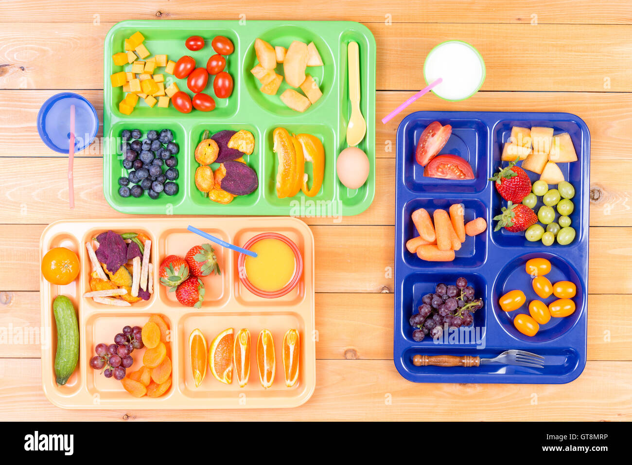 Top down view of colorful plastic lunch trays filled with delicious pieces of fruit and vegetables along side cups of milk or ju Stock Photo