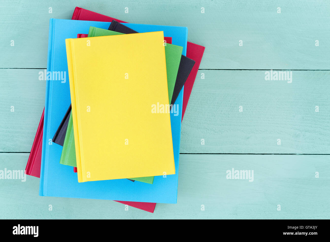Offset stack of multicolored hardcover books piled up on a stained pastel colored green wood table with a bright yellow book on Stock Photo