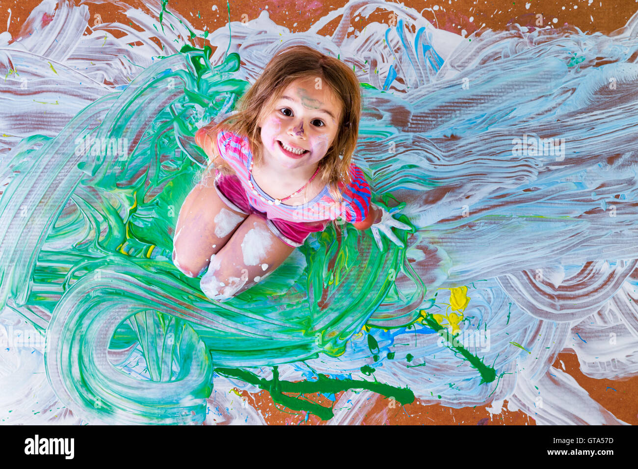 Creative paint splattered little girl having fun with paints kneeling in the center of her artistic modern painting grinning up Stock Photo