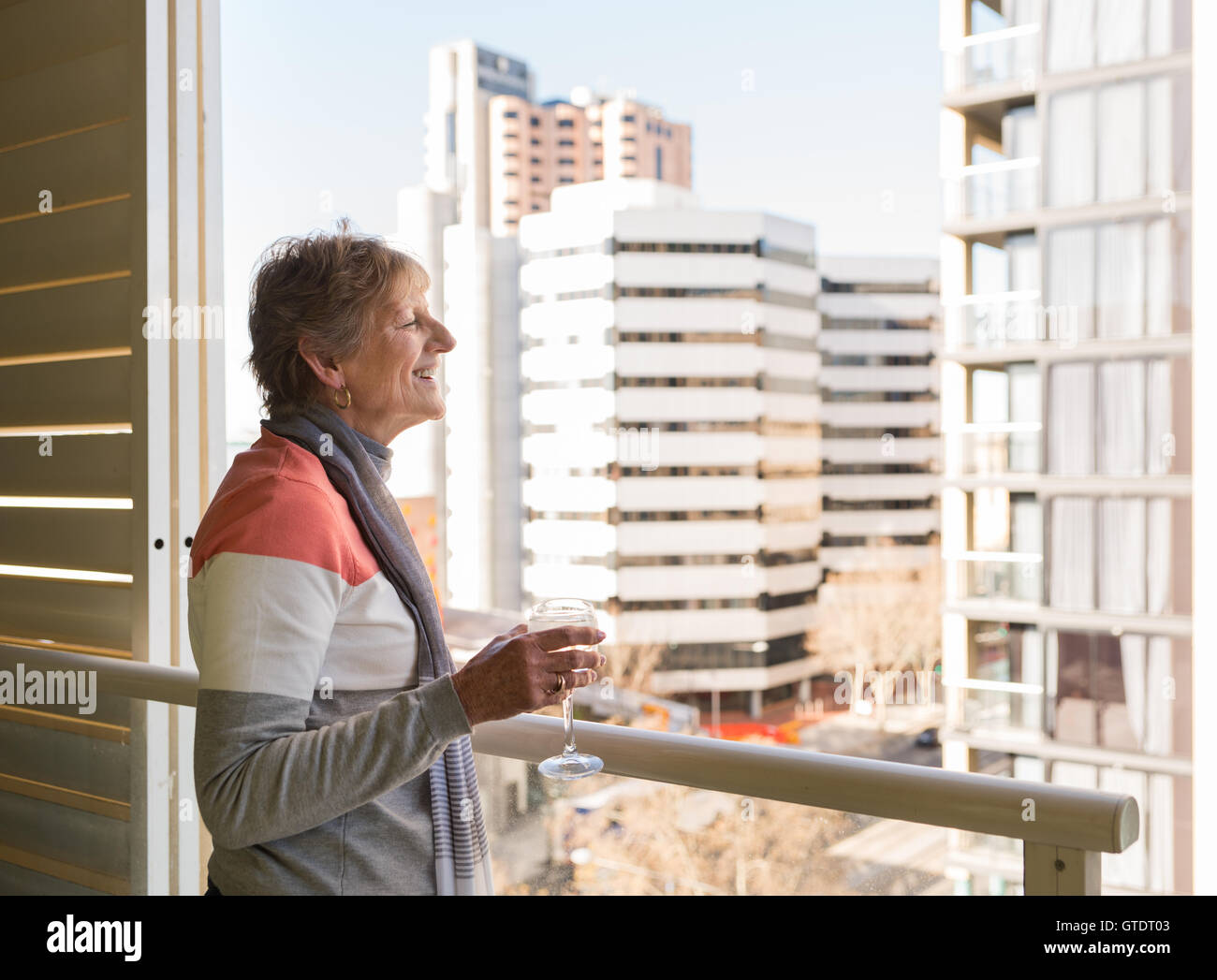 Happy older woman looking at view from highrise apartment holding glass of wine Stock Photo
