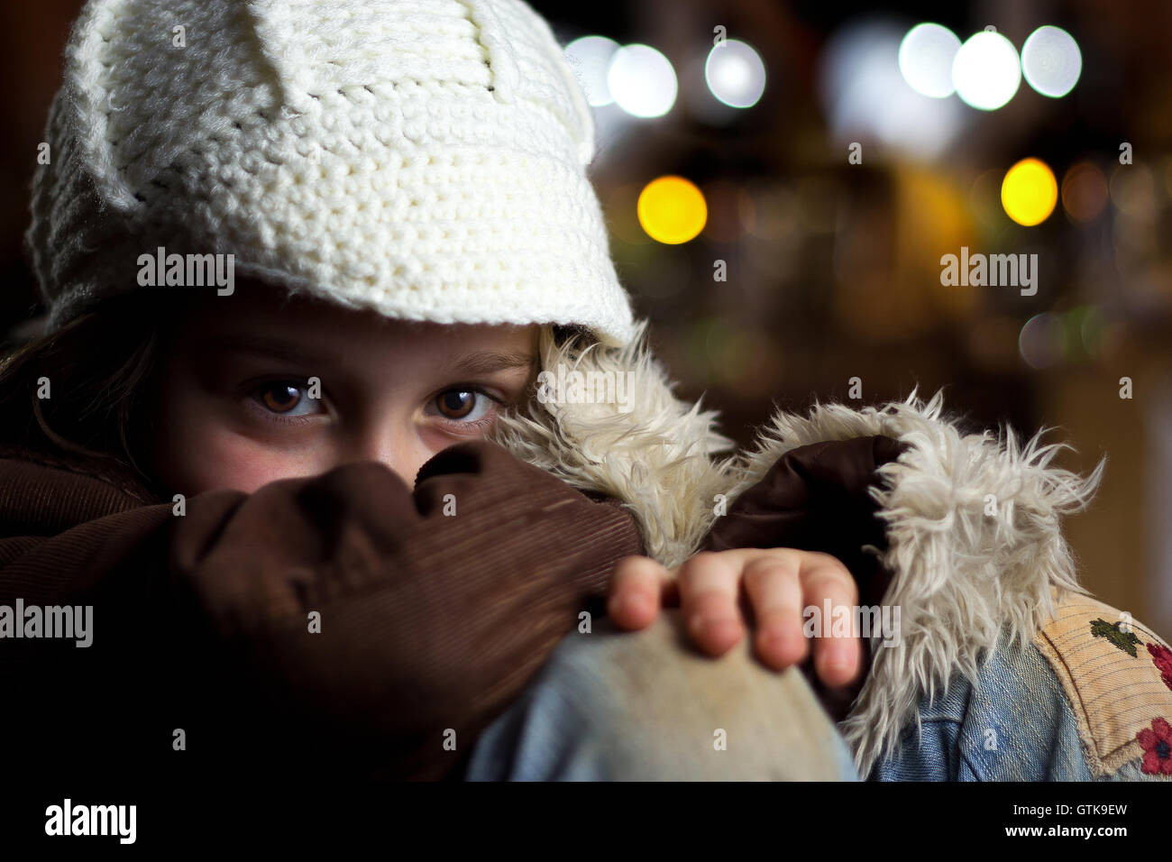 A shy young girl wearing a hat and coat sits in a crouched position looking at the camera. Stock Photo