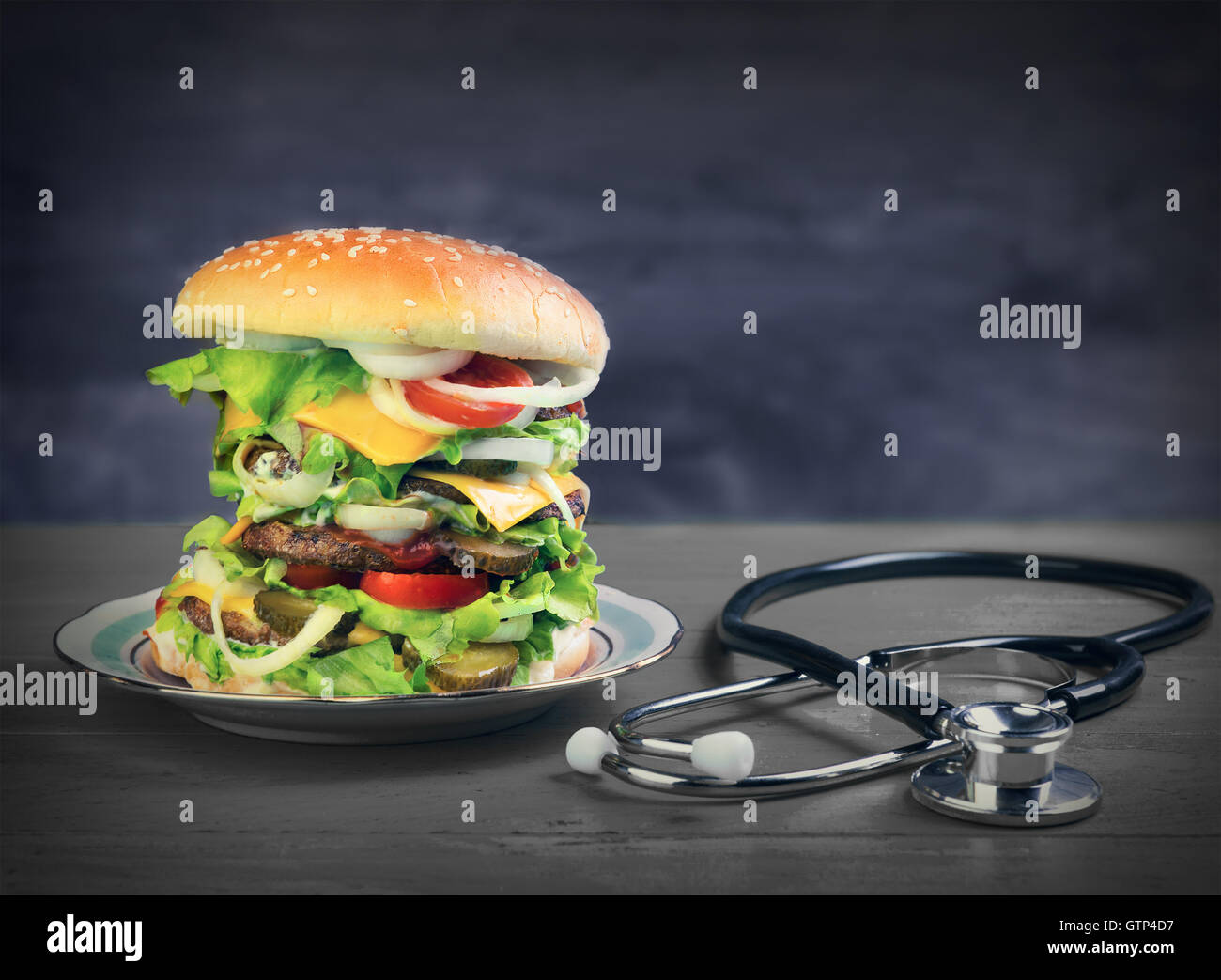 Big cheeseburger deluxe high on green wooden background in rustic style and a stethoscope, unhealthy food Stock Photo