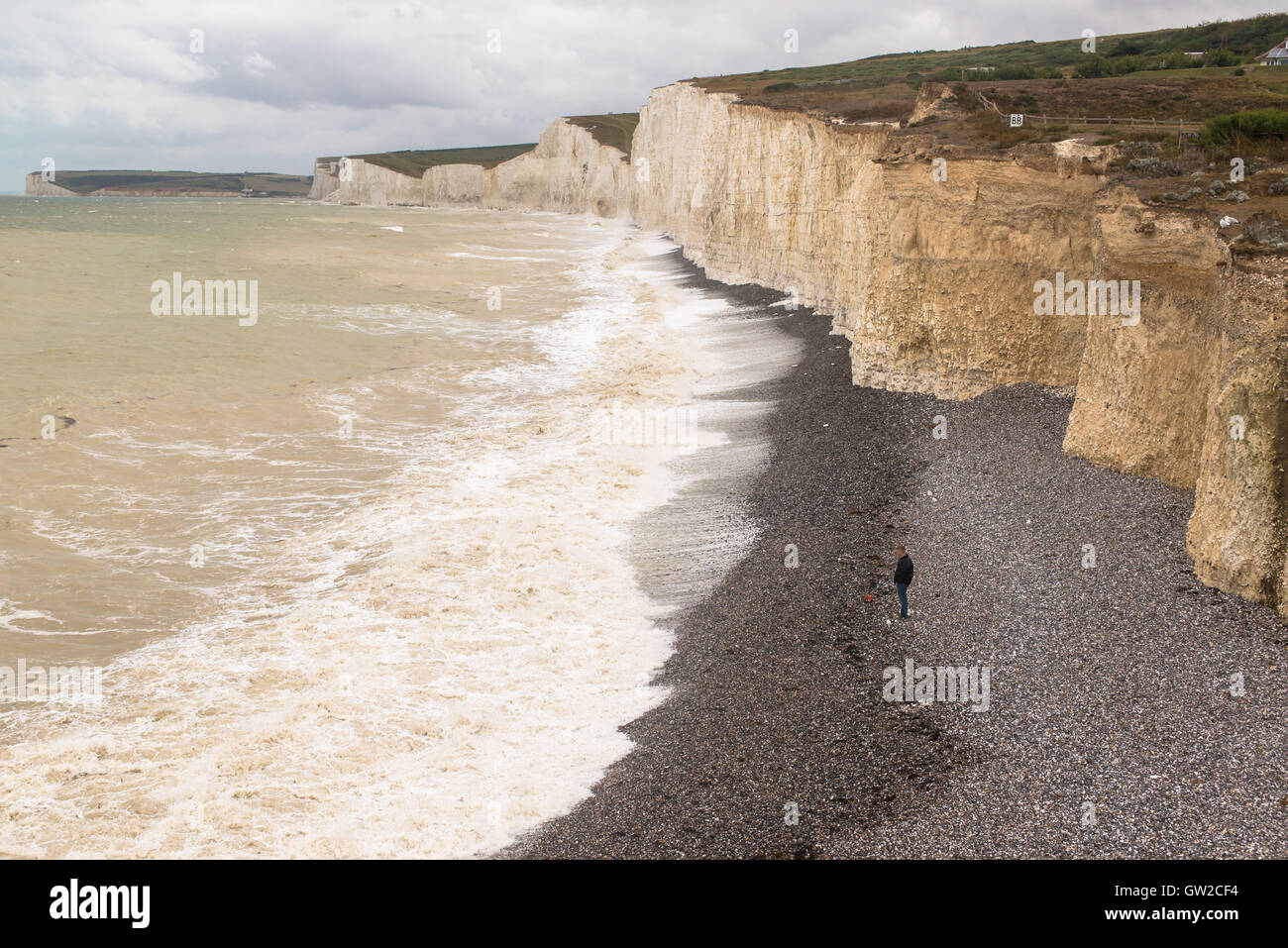 Solitary man standing alone on a gravel beach below the white cliffs of Seven Sisters facing rough sea and waves. Stock Photo