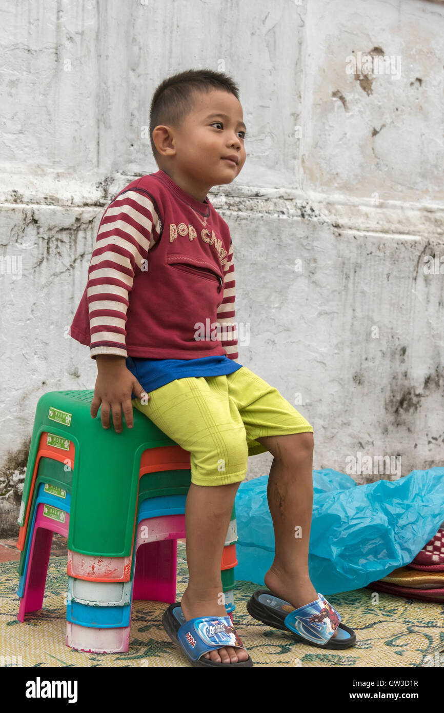 Young boy sitting on a stack of stools at the end of the alms giving ceremony, Wat Sene, Luang Prabang, Laos Stock Photo