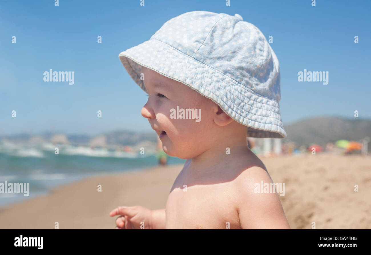 Happy baby boy with blue hat sitting on the sand and observing the seashore on the beach Stock Photo