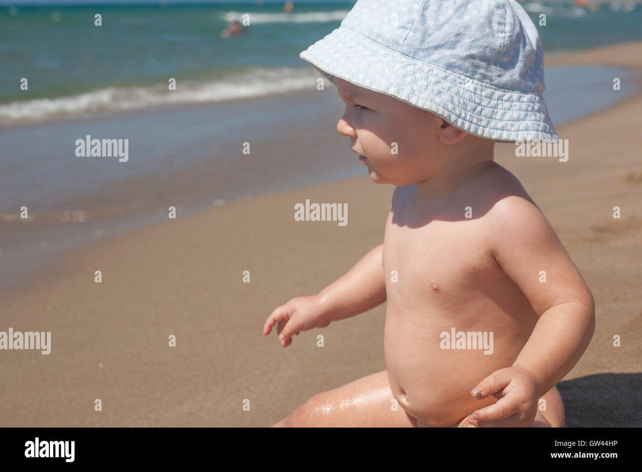 Happy baby boy with blue hat sitting on the sand and observing the seashore on the beach Stock Photo