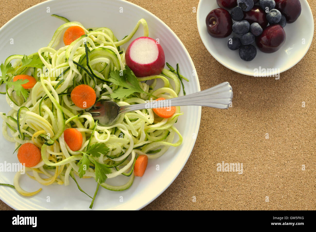 Raw Zucchini noodles with carrots and radish on white plate, over cork background, served with a bowl of blueberries and cherry Stock Photo