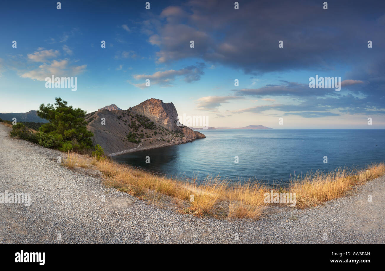 Beautiful panoramic landscape with mountains, path, yellow grass, sea, blue sky and beautiful clouds at sunset in Crimea. Sunset Stock Photo