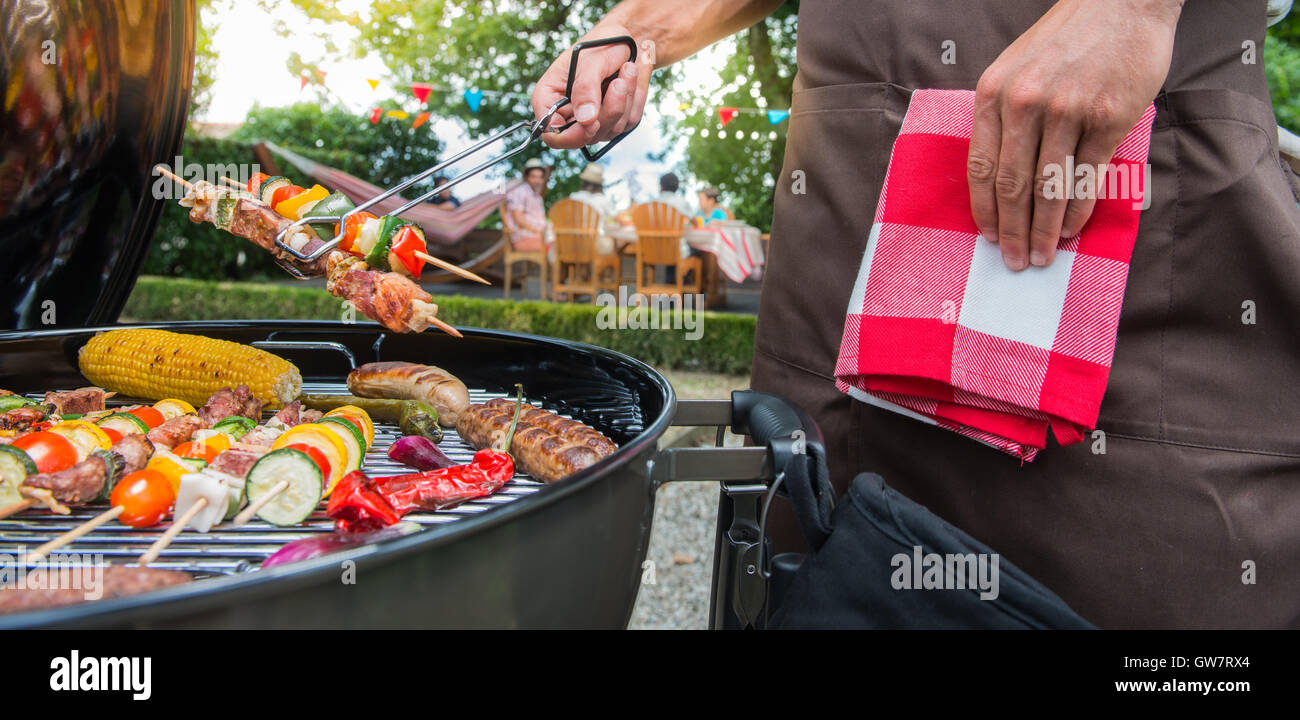 Man grilling meat on garden barbecue party, in the background friends eating and drinking Stock Photo