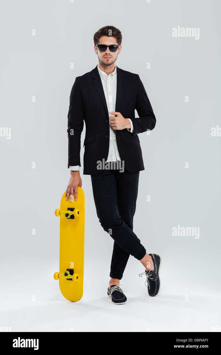Serious young businessman in sunglaasses and suit leaning on skateboard with legs crossed over gray background Stock Photo