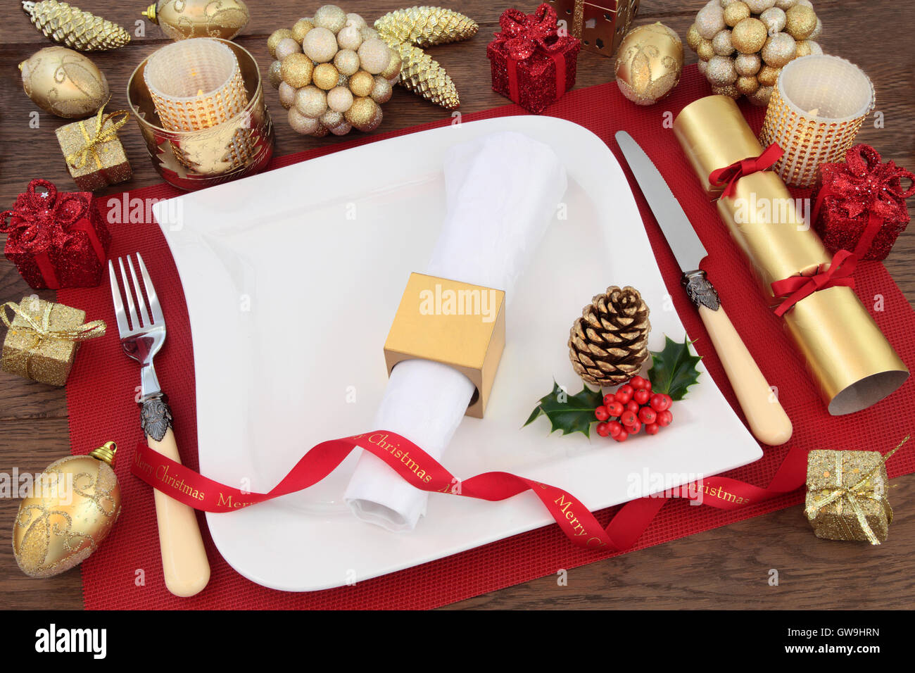 Luxury christmas dinner table setting with white plate, linen serviette, holly, gold bauble decorations, ribbon and cracker. Stock Photo