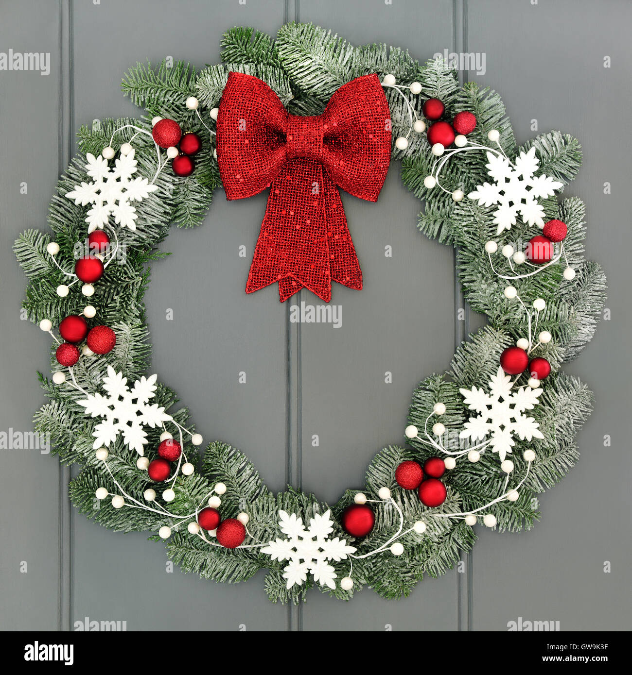 Christmas wreath with red baubles and bow and white snowflake decorations and snow covered blue spruce fir over grey front door Stock Photo