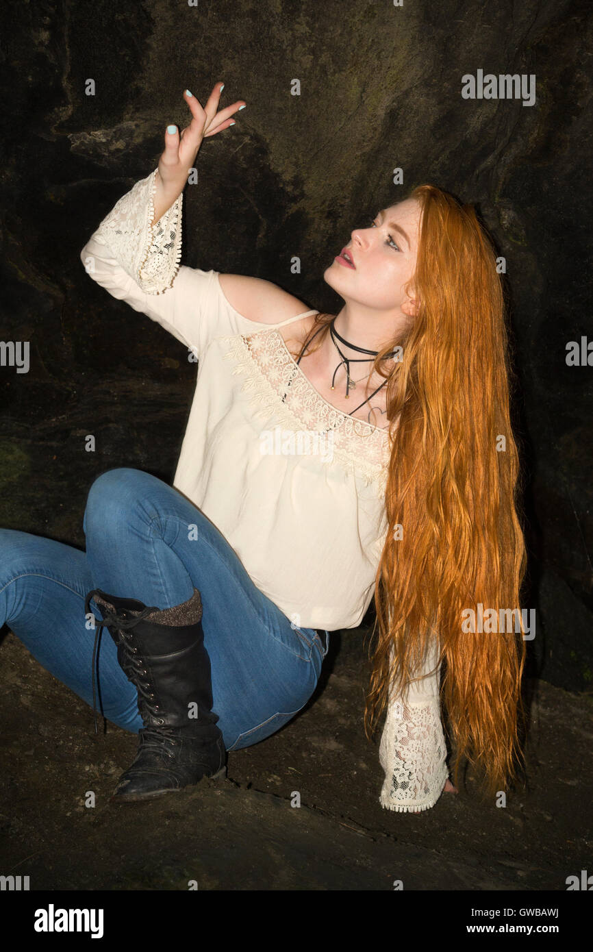 Beautiful red head in skinny jeans, white top and black boots, sitting perilously in the nook of a small cave at an old tunnel. Stock Photo