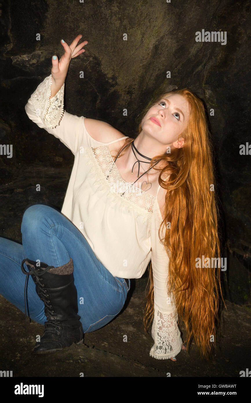 Beautiful red head in skinny jeans, white top and black boots, sitting perilously in the nook of a small cave in Connecticut. Stock Photo
