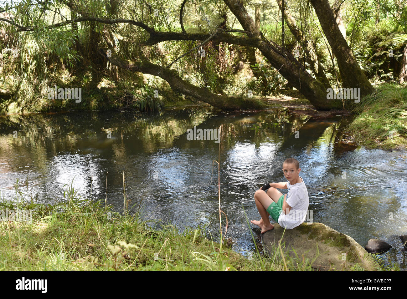 Boy sitting on boulder in small river. Location: Whangarei New Zealand Stock Photo