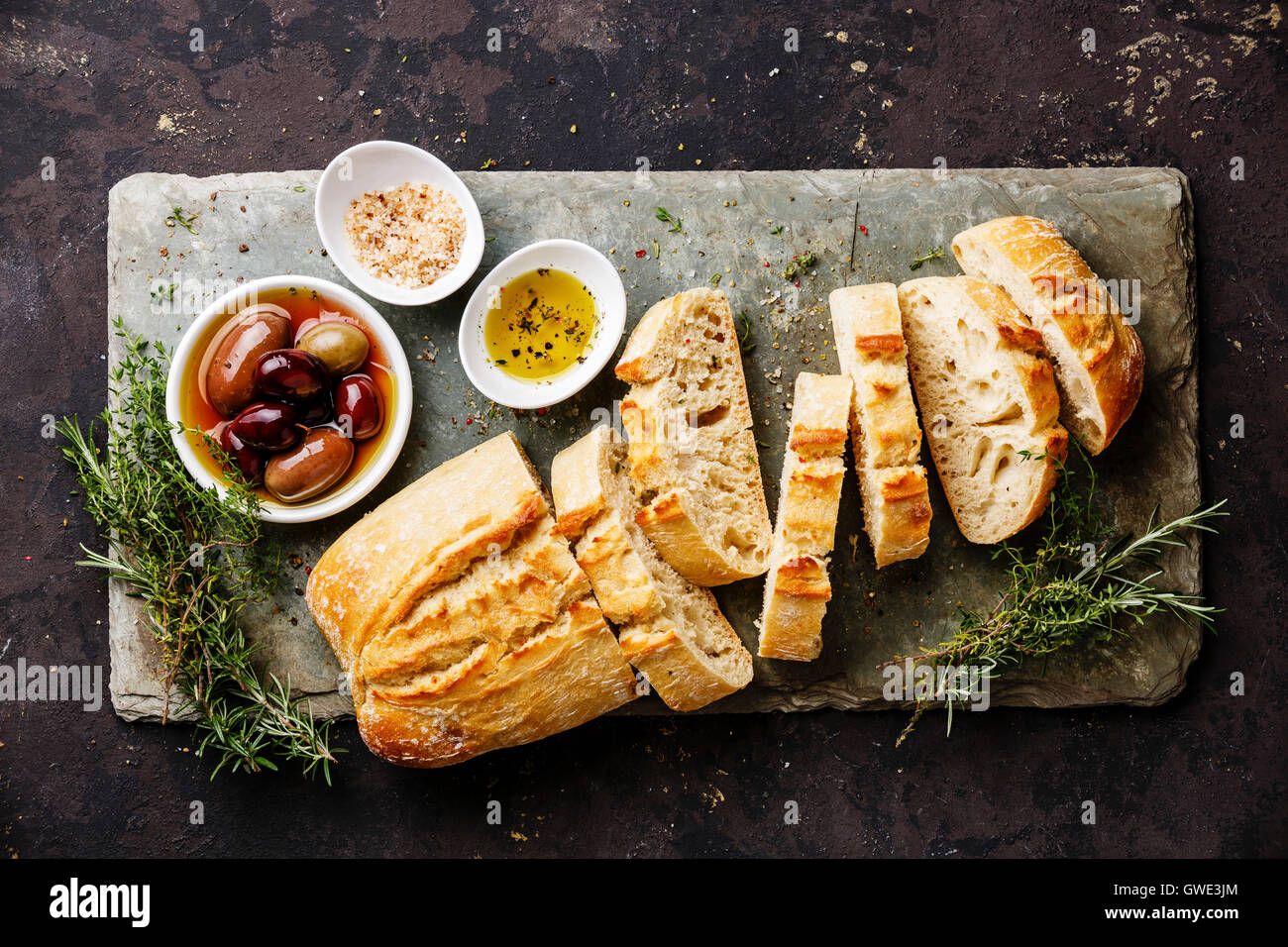 Fresh Ciabatta bread sliced on stone slate board with olives, oil, salt and herbs on dark background Stock Photo