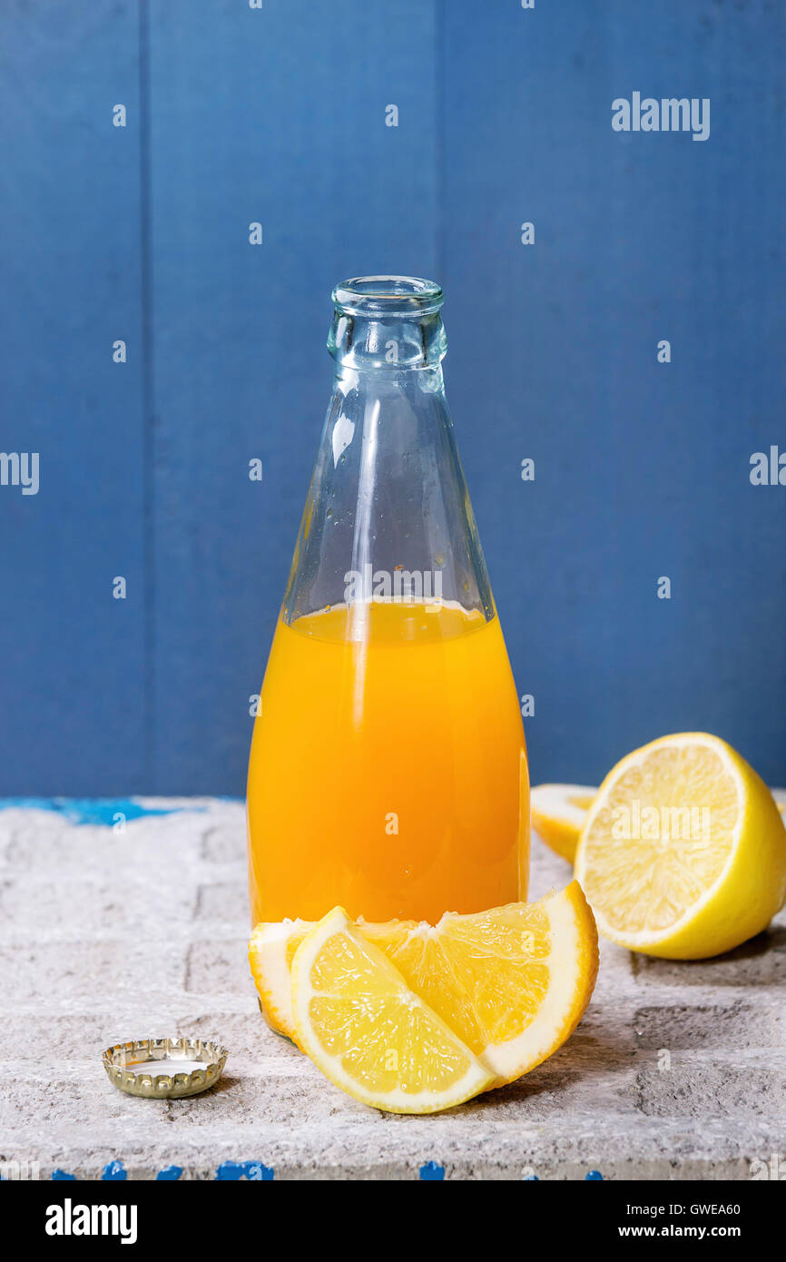 Opened glass bottle of citrus orange and lemon lemonade, standing with sliced fruits on stone board with blue background. With c Stock Photo